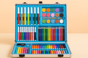 Open art supply case filled with colorful markers, crayons, paint pots, colored pencils, and drawing tools, all arranged neatly in a blue molded tray against a soft peach background.