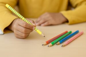 Close-up of a child holding a yellow polka-dotted pencil above a desk, with several colorful dotted pencils lined up on the tabletop in a classroom setting.